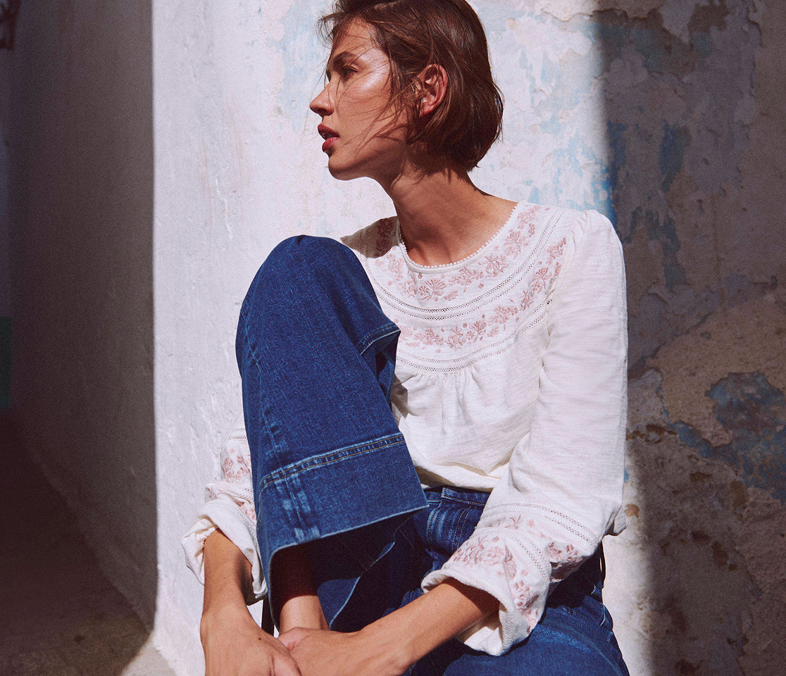 Woman sitting against a textured wall wearing a white lace top and blue jeans.