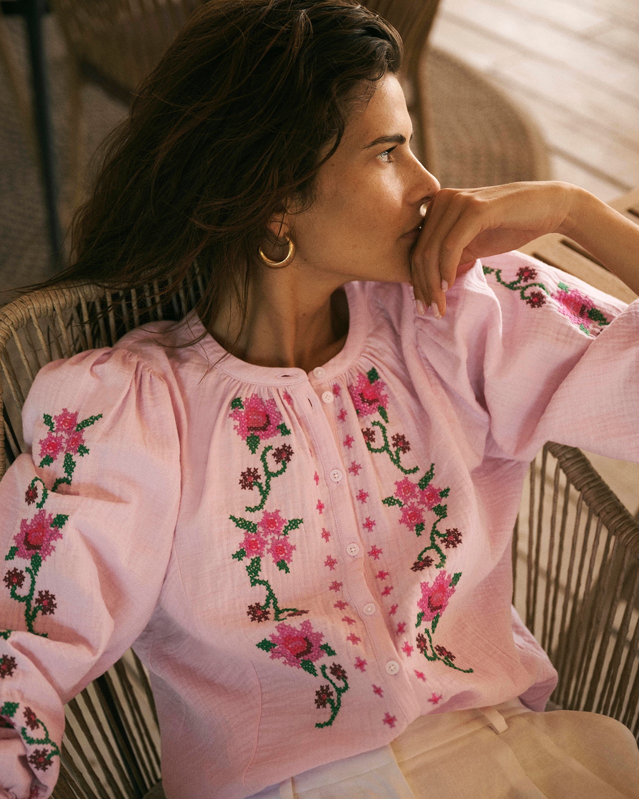 Woman wearing a pink blouse with floral embroidery sitting on a chair.