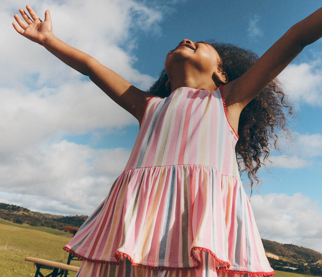 Young girl in a striped dress with arms outstretched against a blue sky.
