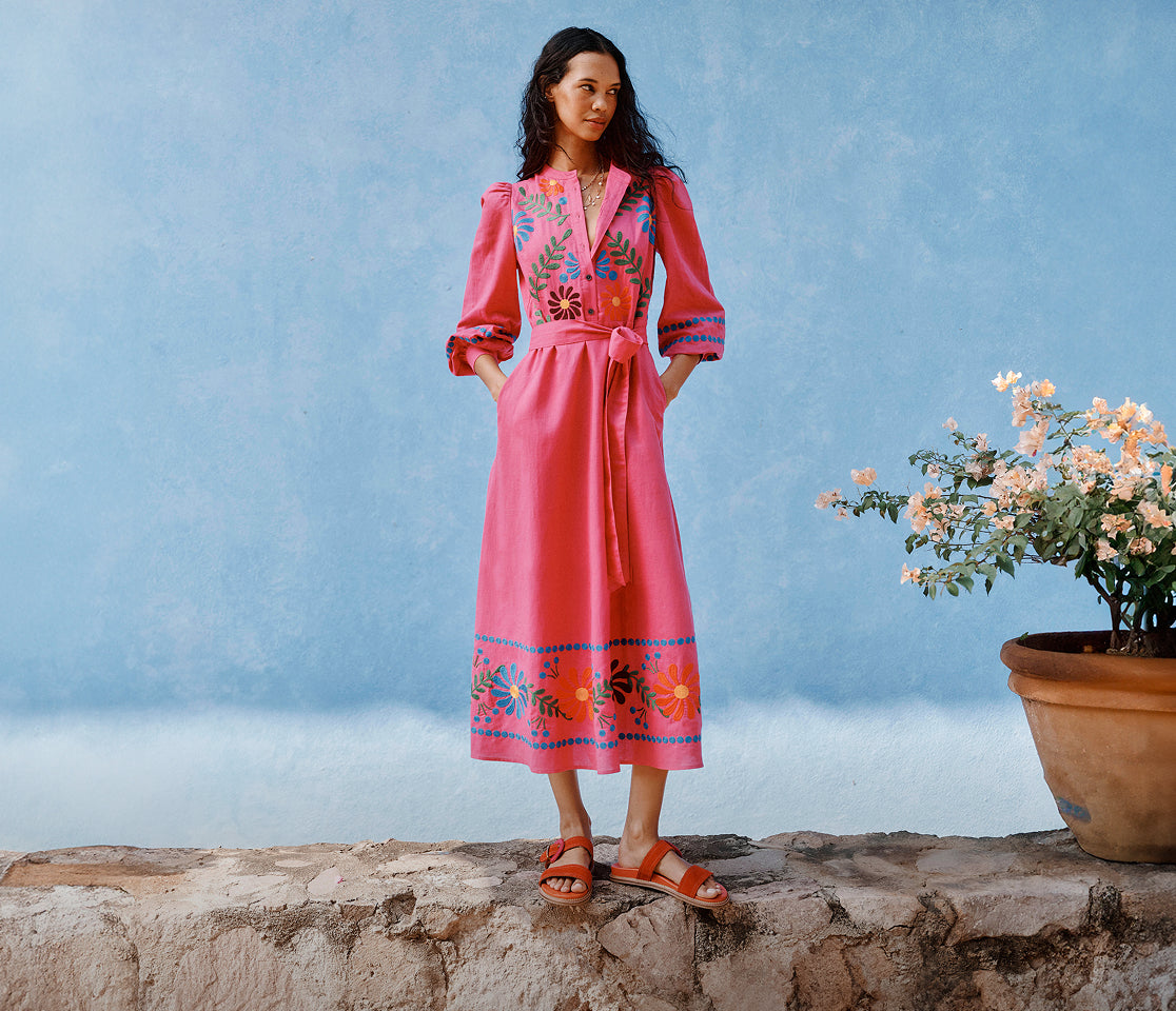 Woman in a pink dress with floral patterns standing against a blue wall with a potted plant.