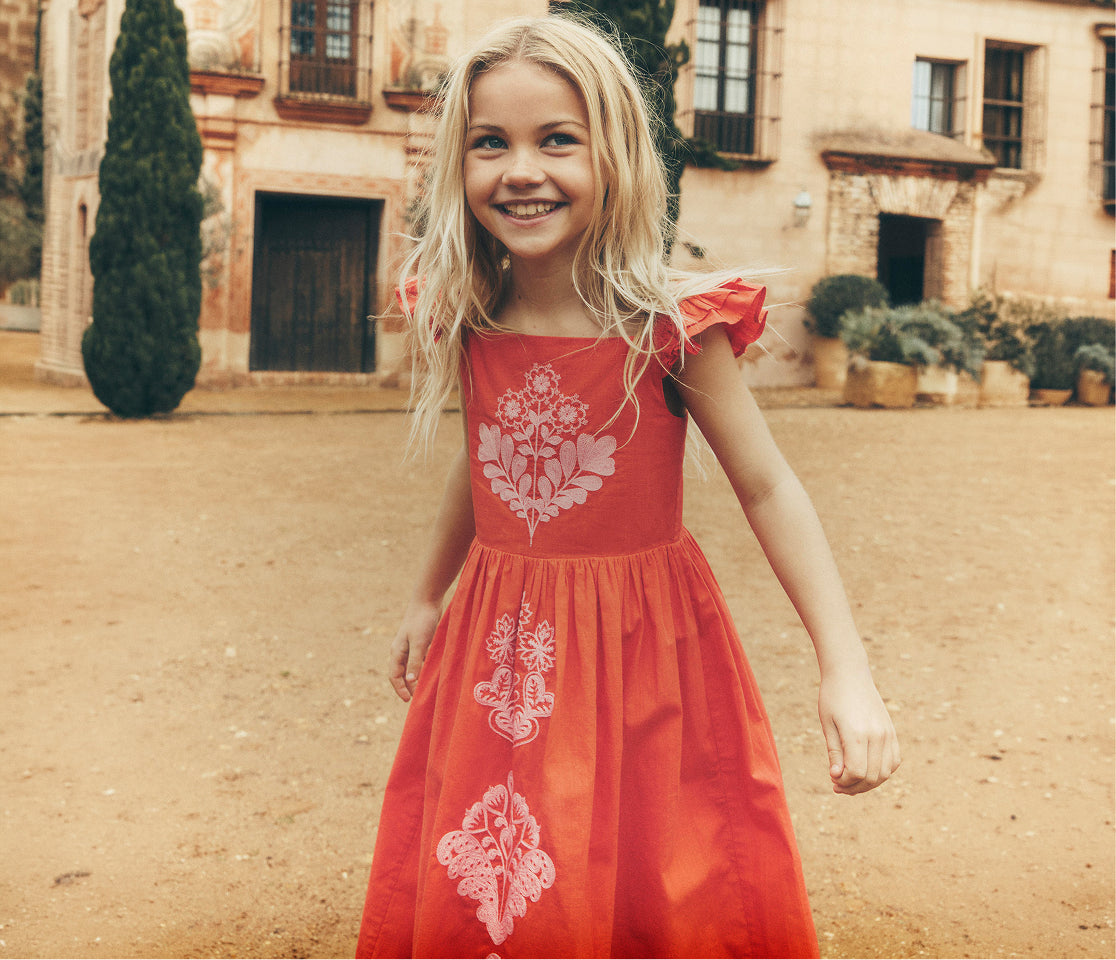 Young girl in a red dress with white floral patterns standing in front of a stone building.