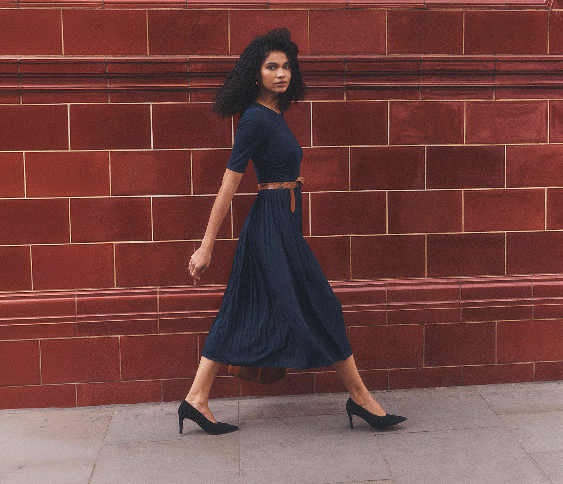 A woman wearing a navy blue midi dress with a thin brown belt and black heels walks in front of a red brick wall.