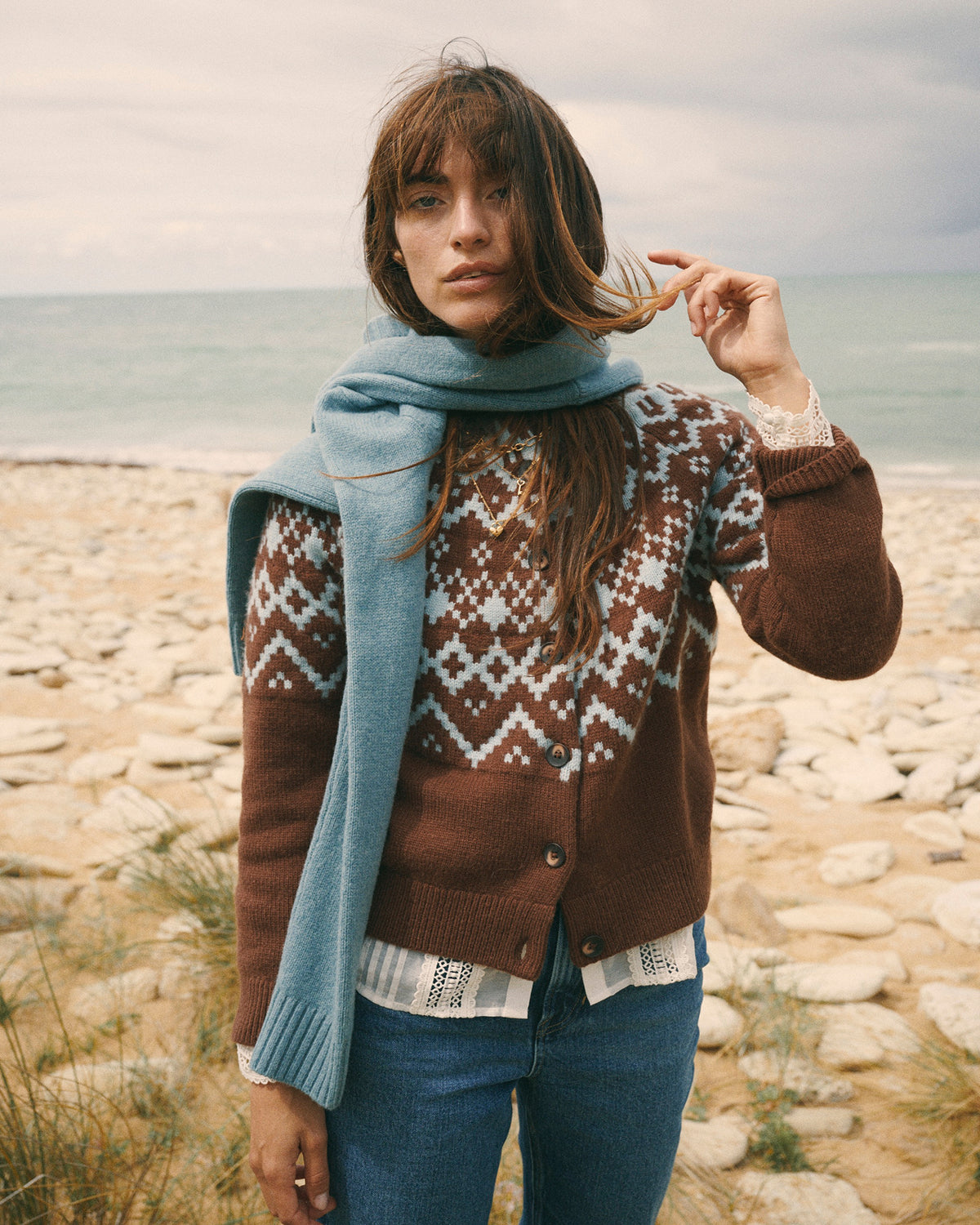 A woman standing on a rocky beach wearing a brown Fair Isle knit cardigan, a light blue knit scarf, and blue jeans, with the ocean and cloudy sky in the background.