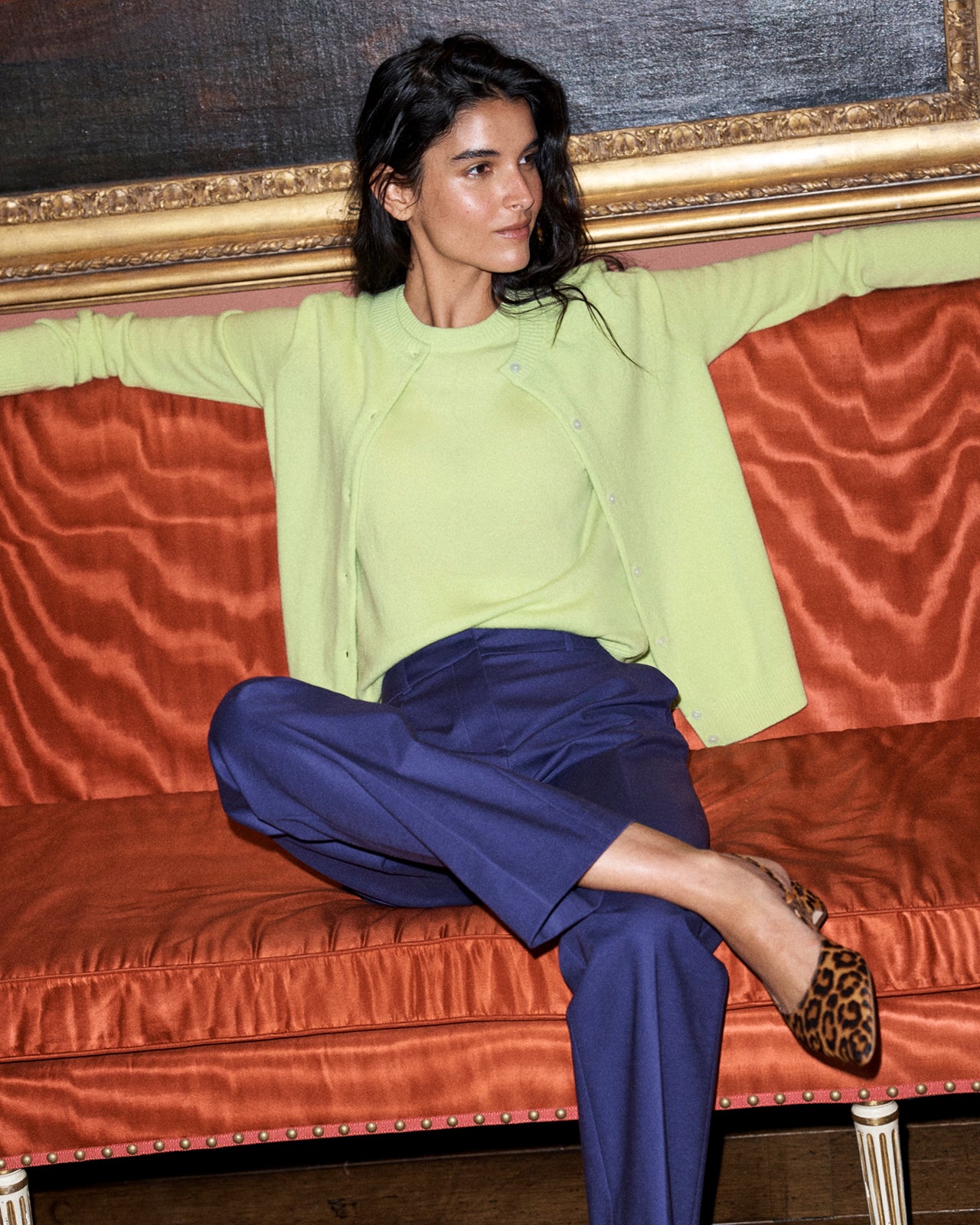 A woman sits on an orange velvet sofa wearing a light green cardigan and top with navy blue trousers, and leopard-print shoes.