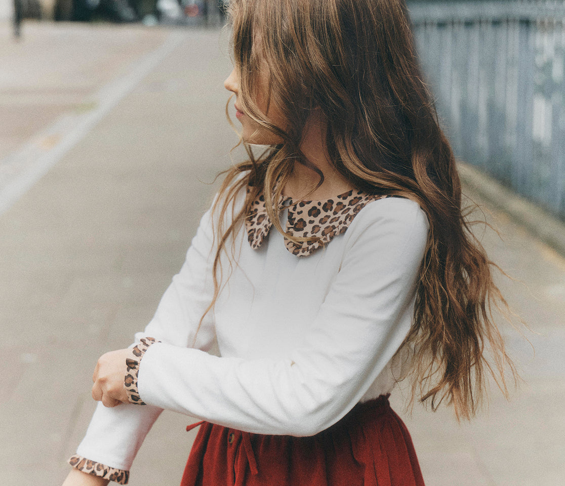 A young girl with long brown hair is shown from the side and back, wearing a white long-sleeve top with a brown and beige leopard-print collar and matching cuffs, paired with a dark red skirt, set against an outdoor blurred background.