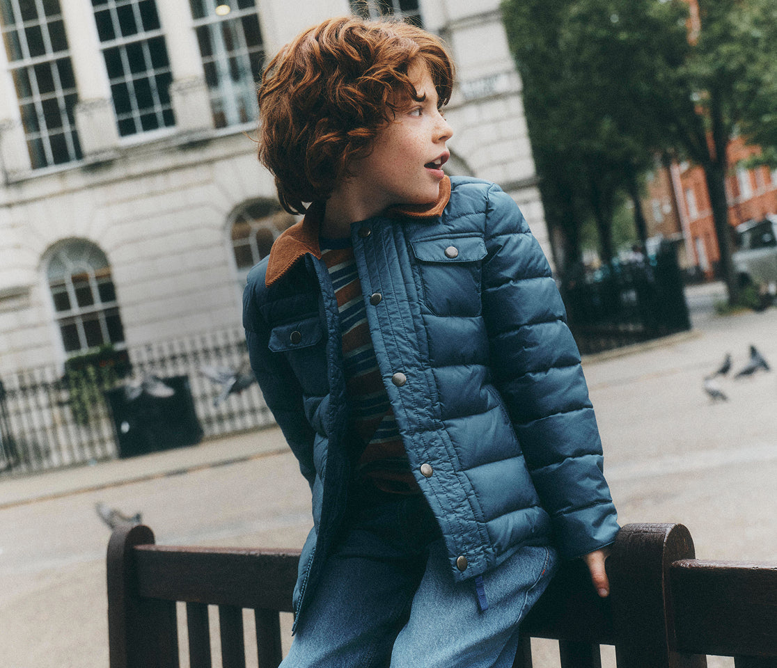 A child with curly red hair wearing a blue puffer jacket and blue jeans, leaning on a dark wooden bench in an outdoor urban setting with a building and trees in the background.