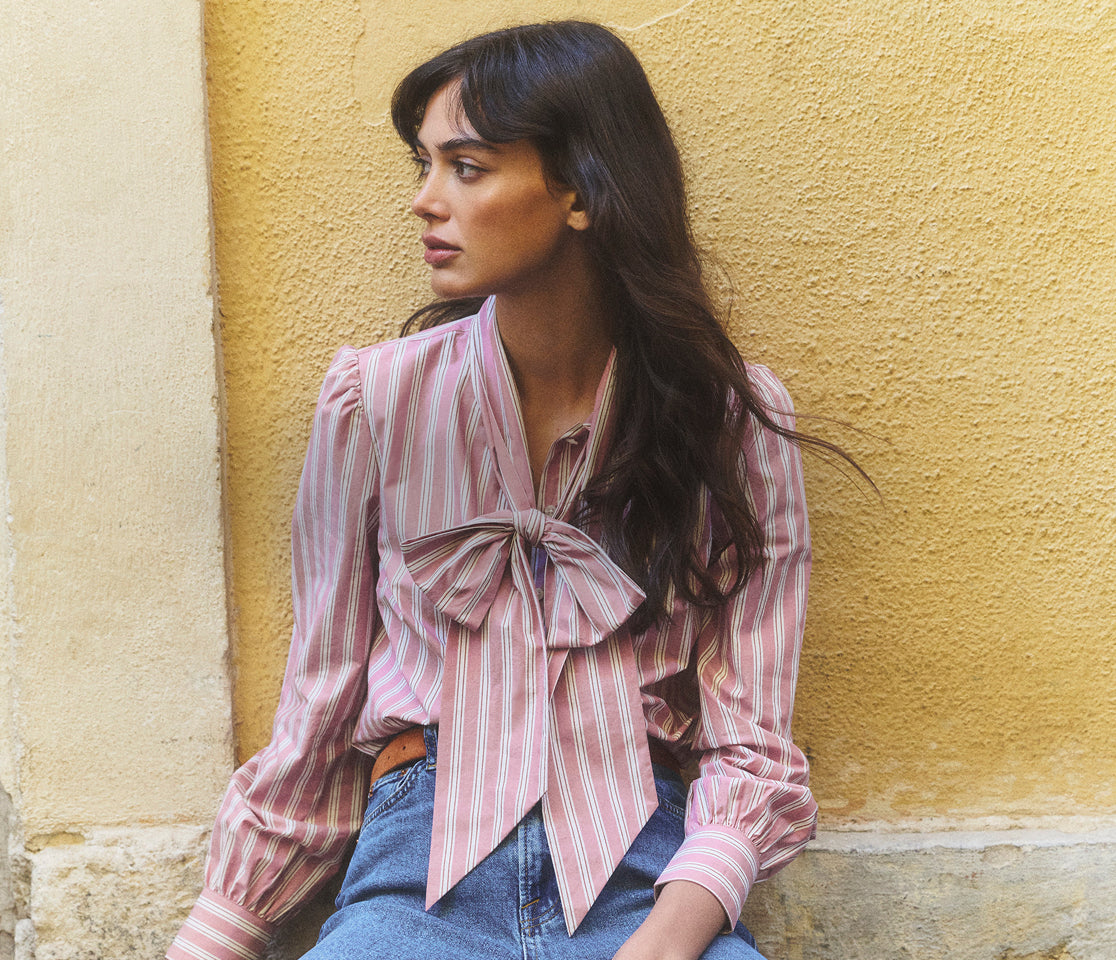 Woman wearing a pink striped blouse with a bow tie, sitting against a yellow wall.