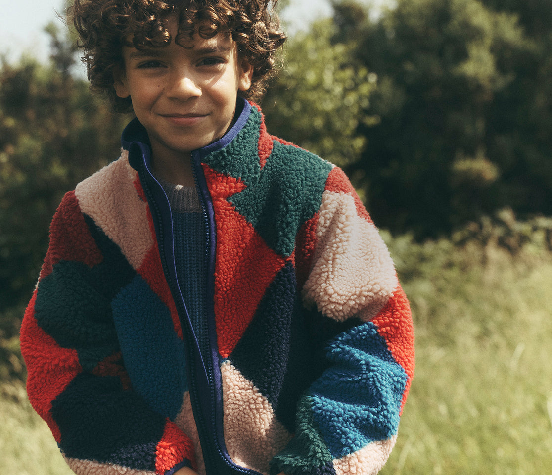 Child standing in a sunlit grassy field wearing a plush geometric fleece jacket in red, dark green, navy, blue and cream over a knitted jumper and blue jeans. The jacket has a high collar, front zip and contrasting colour-block pattern, with leafy trees softly blurred in the background.