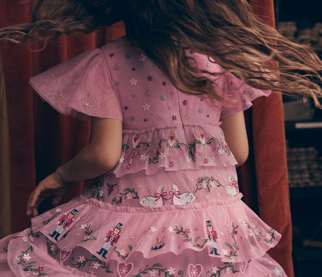 A child wearing a pink tulle dress with embroidered floral and nutcracker patterns, shown from behind with long brown hair blowing in the wind, set against a dark background with red drapery.