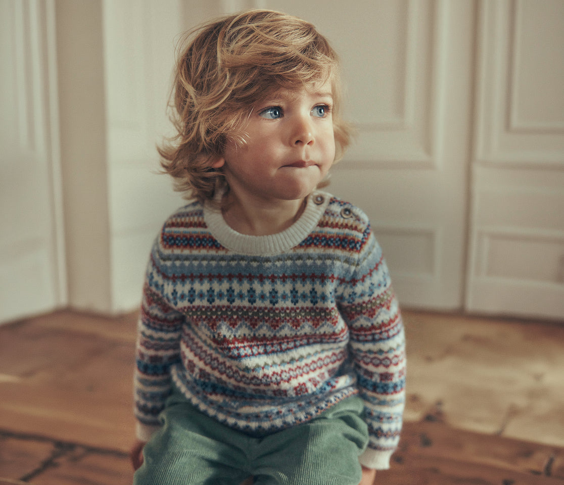 A toddler with wavy light brown hair sits on a wooden floor in front of white paneled doors, wearing a long-sleeve knit sweater with a multicolor Fair Isle pattern and green pants.