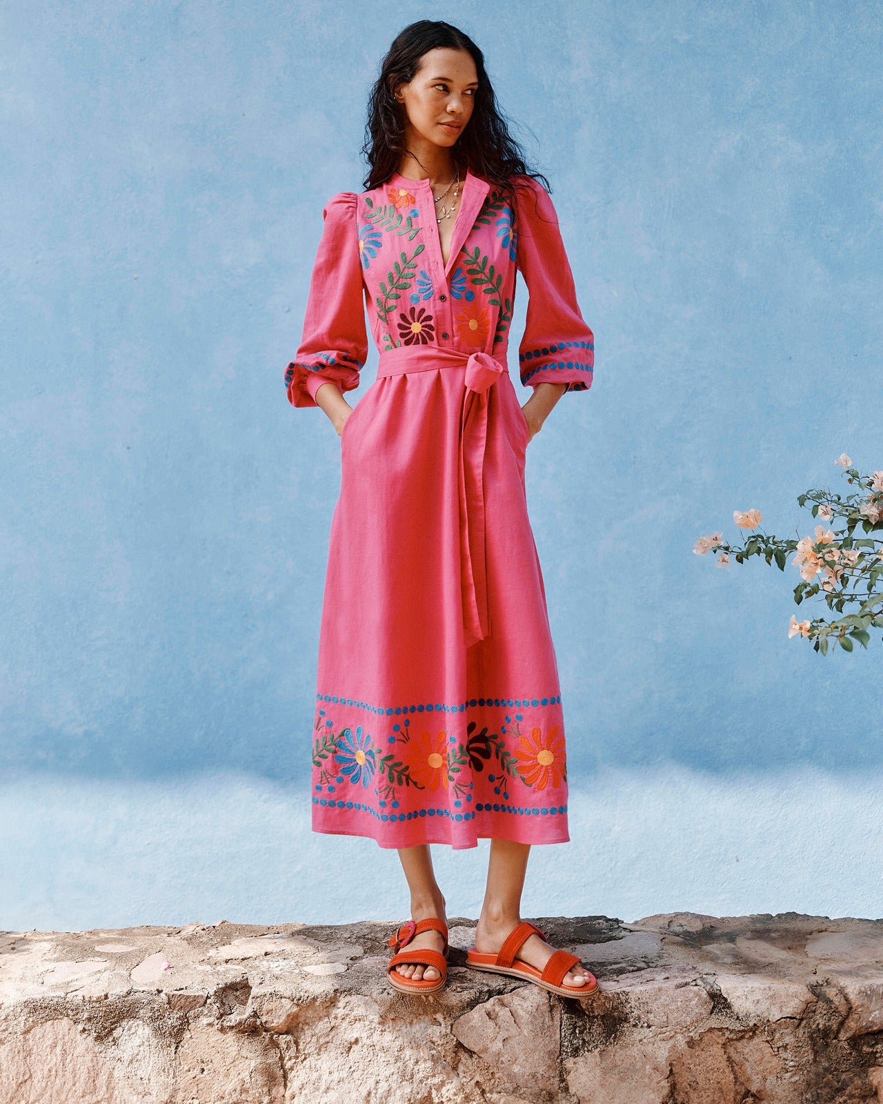 Woman wearing a pink dress with floral embroidery standing on a rock against a blue sky.