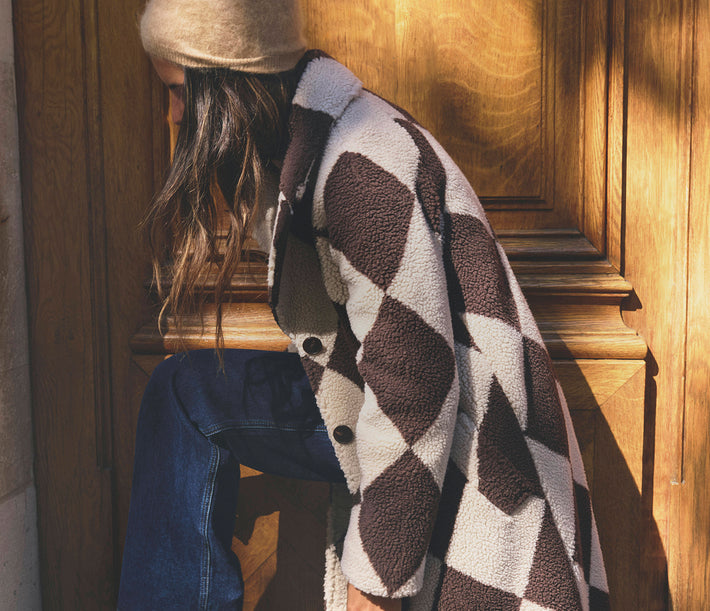 A person wearing a brown and cream argyle sherpa fleece jacket, blue jeans, and a beige beanie sits against a warm wooden door in direct sunlight.