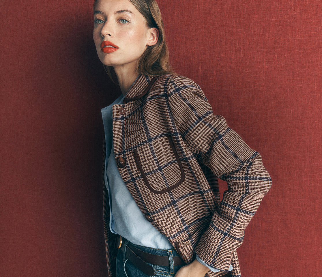 A woman wearing a brown and beige plaid blazer over a light blue shirt and dark jeans, posing against a textured dark red wall.