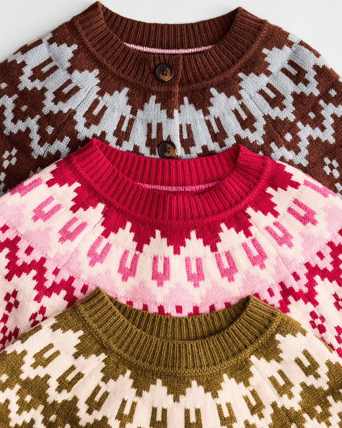 Three knit crewneck sweaters with Fair Isle patterns, stacked diagonally. The top sweater is brown and light gray, the middle is pink and white, and the bottom is olive green and cream.
