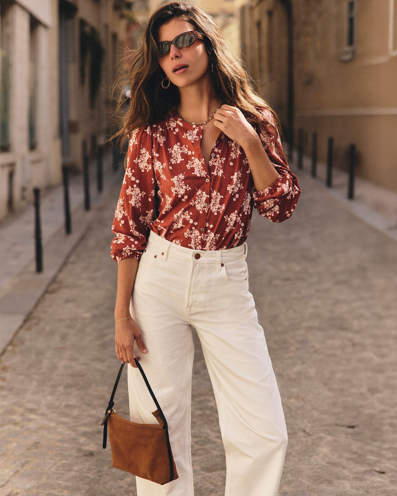 A woman stands on a sunlit cobblestone street wearing a rust-red floral blouse with white sleeves, white wide-leg jeans, dark sunglasses, and holding a brown handbag.