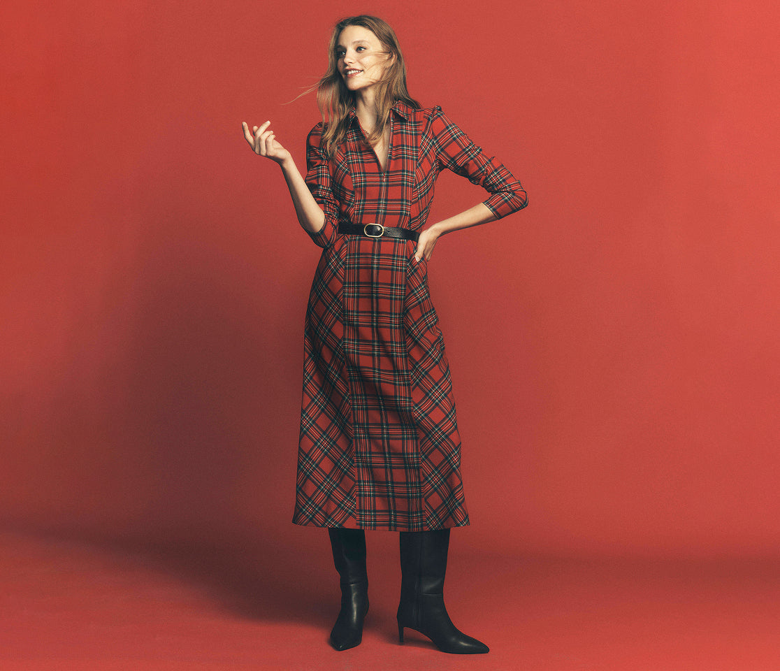 A woman modeling a red and black plaid midi shirt dress with a black belt and black knee-high boots, standing against a solid red studio background.