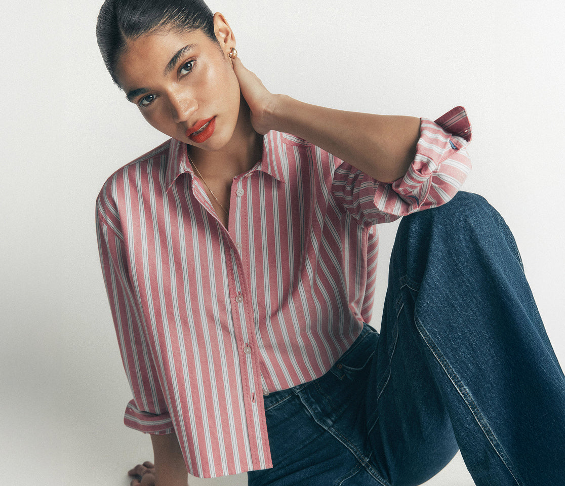 A woman wearing a pink and white vertically striped button-down shirt with rolled-up sleeves and dark blue jeans, posing against a light gray studio background.