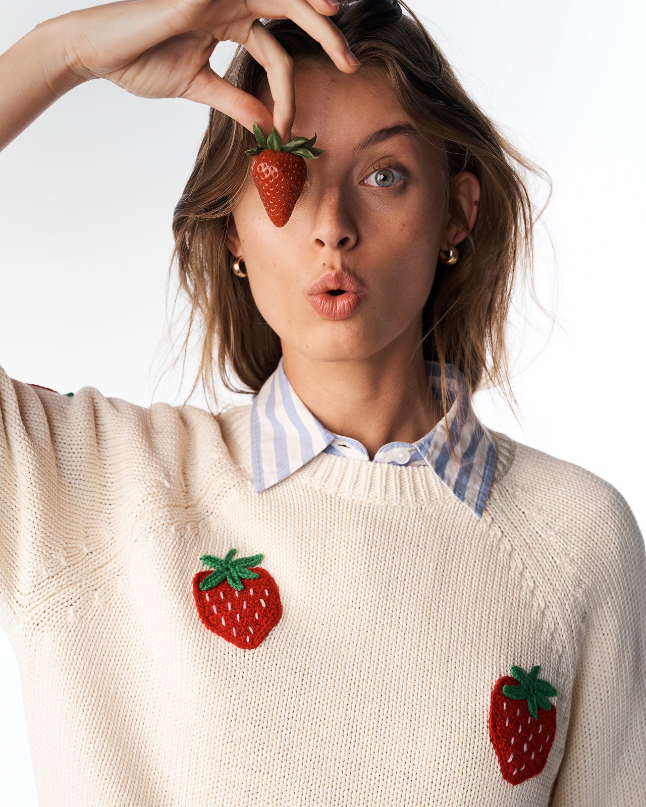 Woman wearing a sweater with strawberry designs, holding a strawberry to her eye.