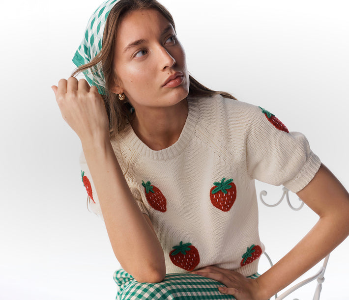 Woman wearing a beige sweater with strawberry patterns and a green checkered skirt.