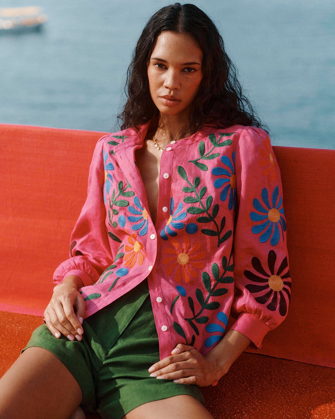 Woman wearing a pink embroidered jacket with floral patterns, sitting on a red surface with water in the background.