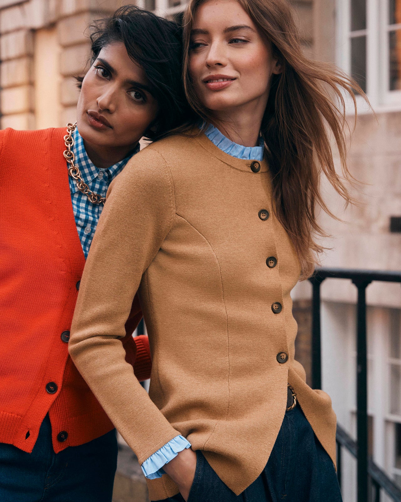 Two women standing close together, one in a red cardigan and blue checkered shirt, the other in a brown cardigan with buttons.