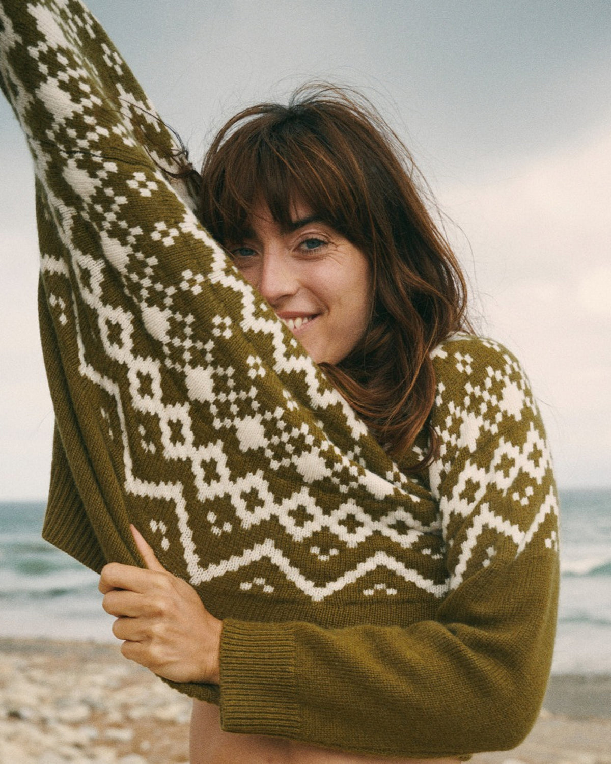 A woman on a beach wearing an olive green and white geometric patterned knit sweater, holding the sleeve up to show the pattern.