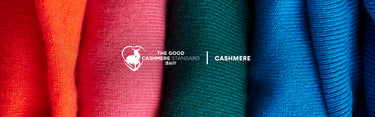Four vertical rolls of textured cashmere fabric in orange, red, green, and blue, with a white 'The Good Cashmere Standard | Cashmere' logo centered over the middle rolls.