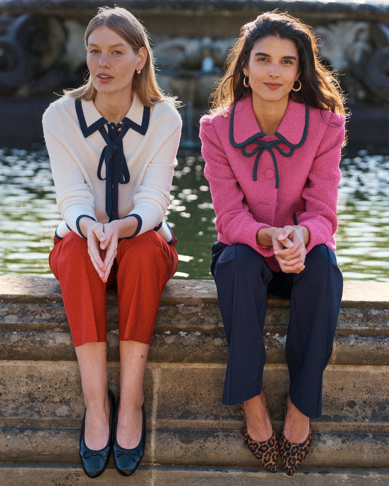 Two women sitting on stone steps in front of a fountain, wearing matching coordinated outfits. The woman on the left wears a cream blouse with a dark navy collar and bow tie detail, paired with rust-red cropped pants and dark navy flats.