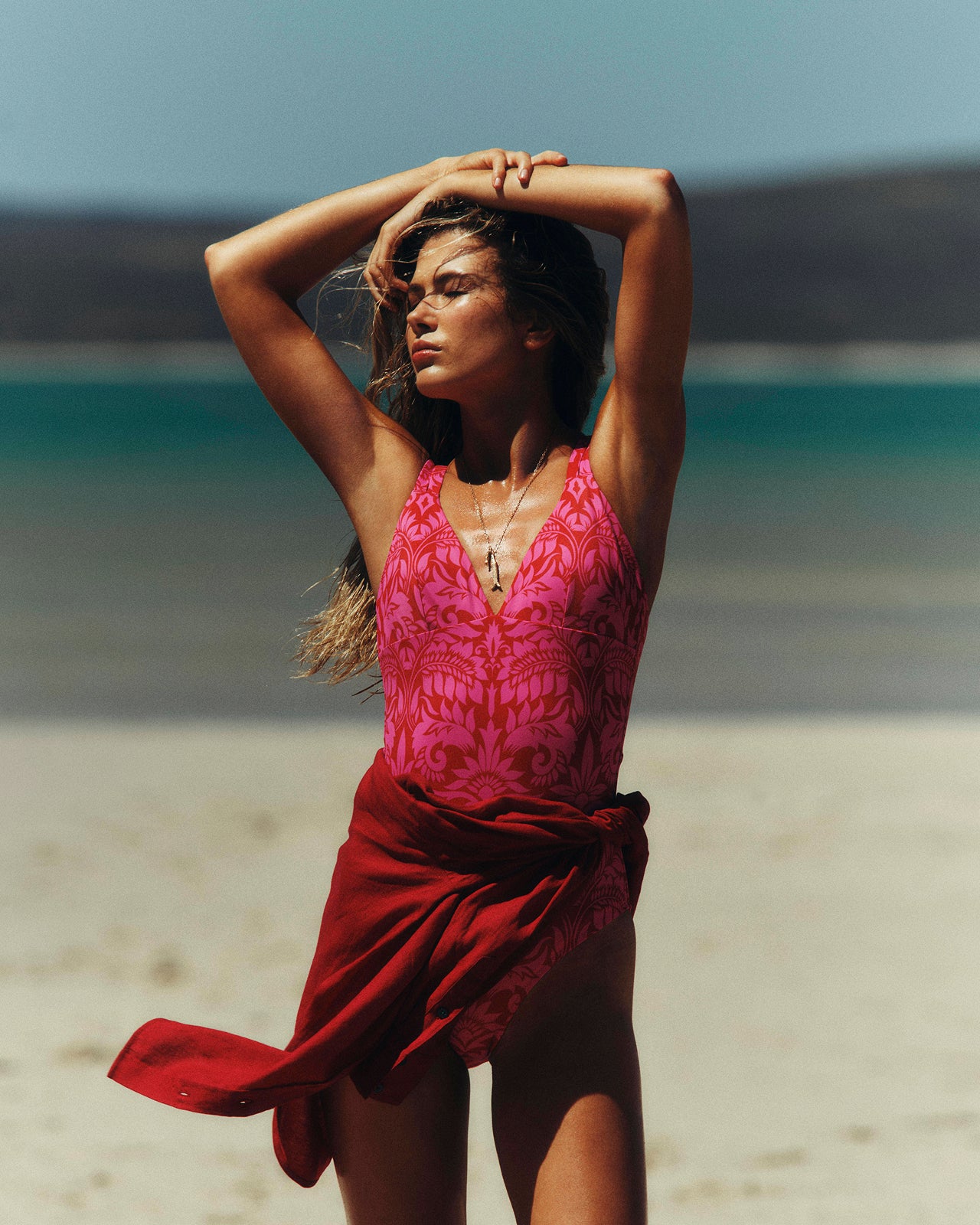 Woman in a pink top and red skirt standing on a beach with turquoise water and blue sky.