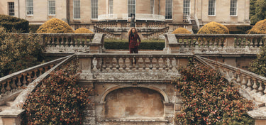 A woman in a dark red coat stands on a stone terrace in front of a grand classical mansion with manicured gardens and stone balustrades.