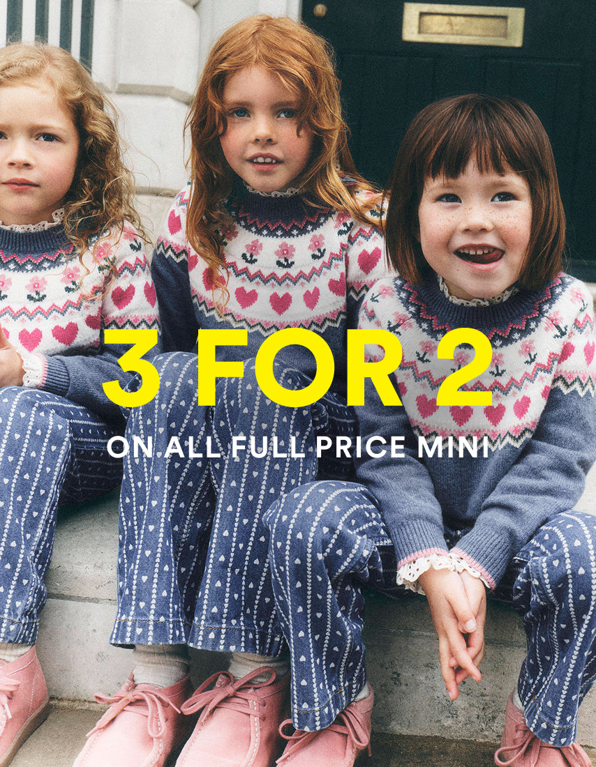 Three young girls sitting on stone steps wearing matching knit sweaters and trousers with a heart and star pattern. Large yellow text across the center reads '3 FOR 2' with smaller white text below reading 'ON ALL FULL PRICE MINI'.