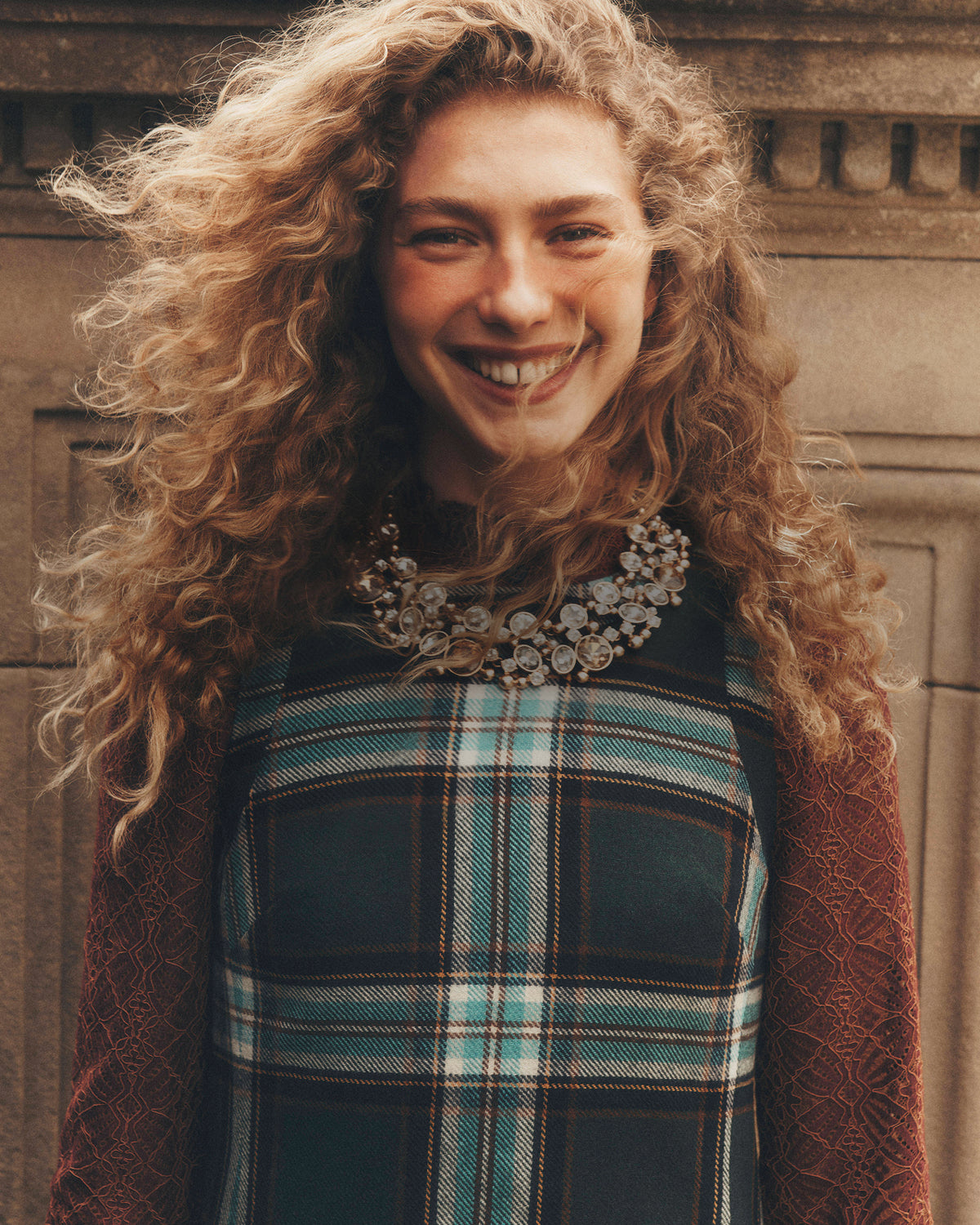 A smiling woman with voluminous curly light brown hair wears a dark teal and brown plaid dress with a brown textured cardigan and a large floral statement necklace, set against a stone wall background.