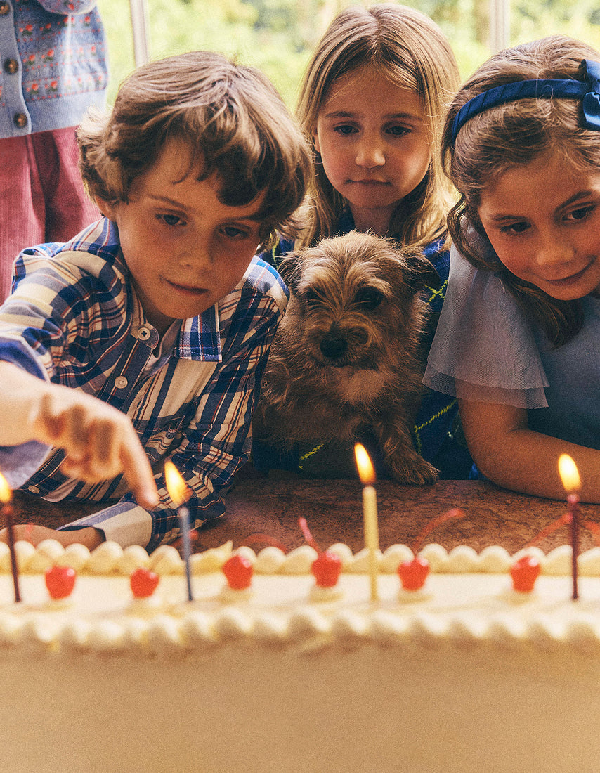 Three children and a small dog gathered around a birthday cake with lit candles in a warm indoor setting.