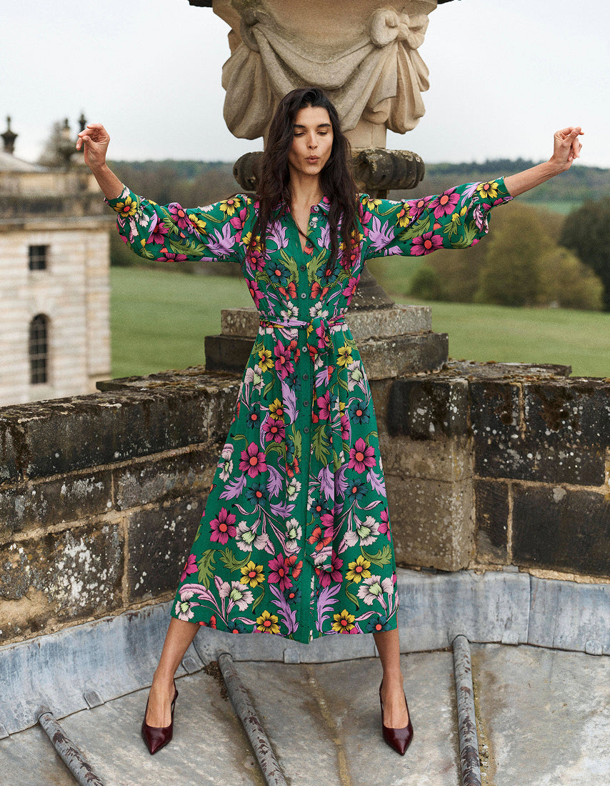 A woman stands outdoors on a stone terrace wearing a long-sleeved midi dress with a vibrant green base and a colorful floral print in pink, yellow, and purple. She is posing with her arms outstretched, with a stone statue and a country estate in the background.