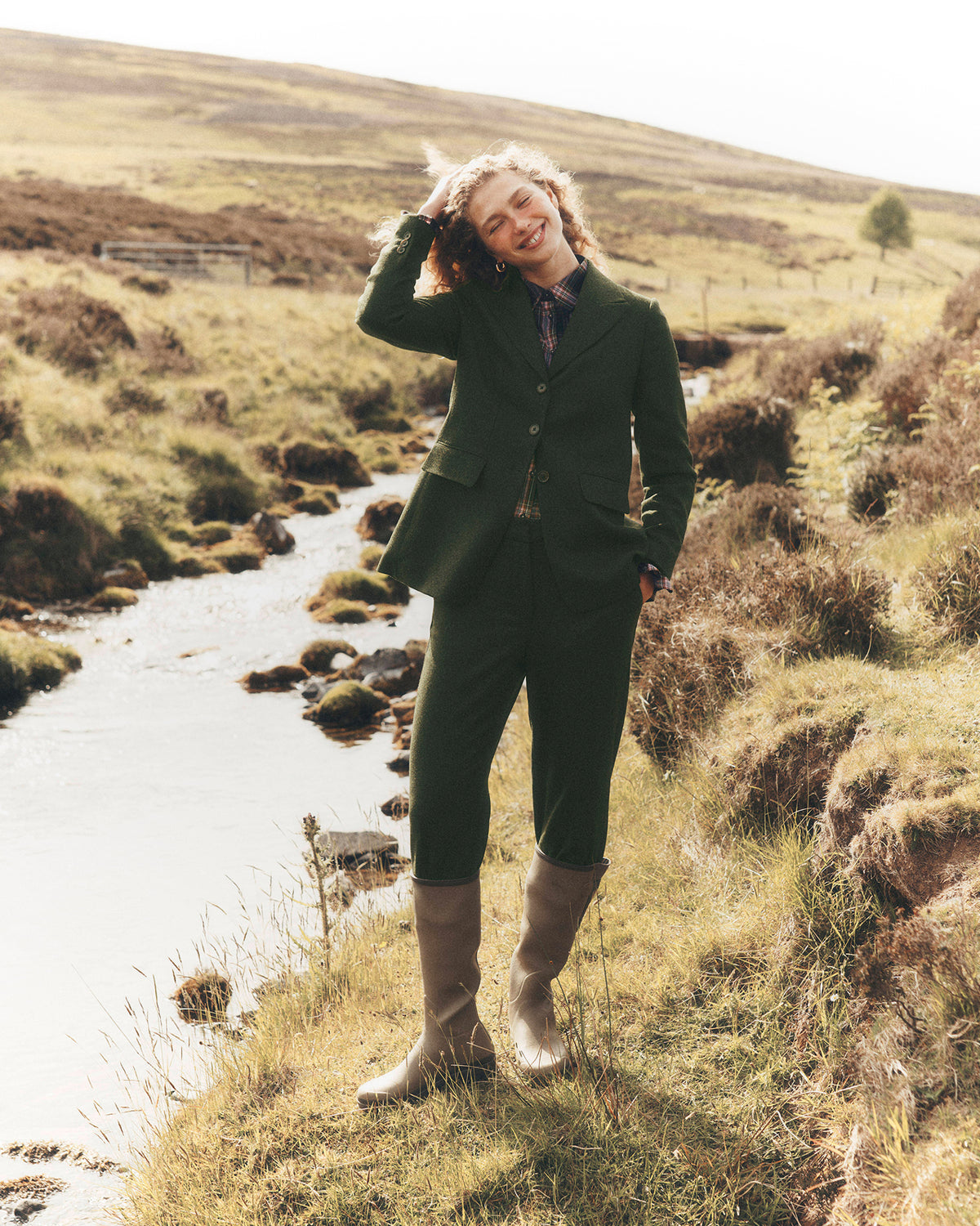 A woman stands outdoors in a grassy, hilly landscape next to a small stream, wearing a dark green tailored suit with cropped trousers, a patterned shirt, and tall light brown rain boots.