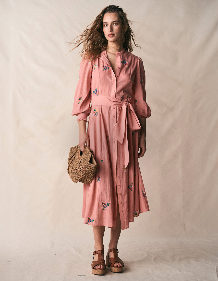 Woman wearing a pink dress with floral patterns, holding a woven bag against a plain background