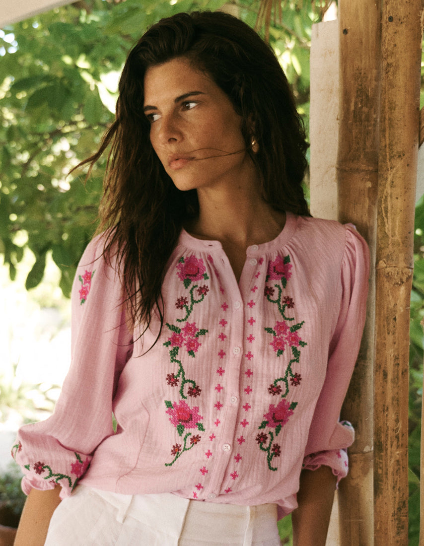 Woman wearing a pink embroidered blouse standing outdoors with greenery in the background