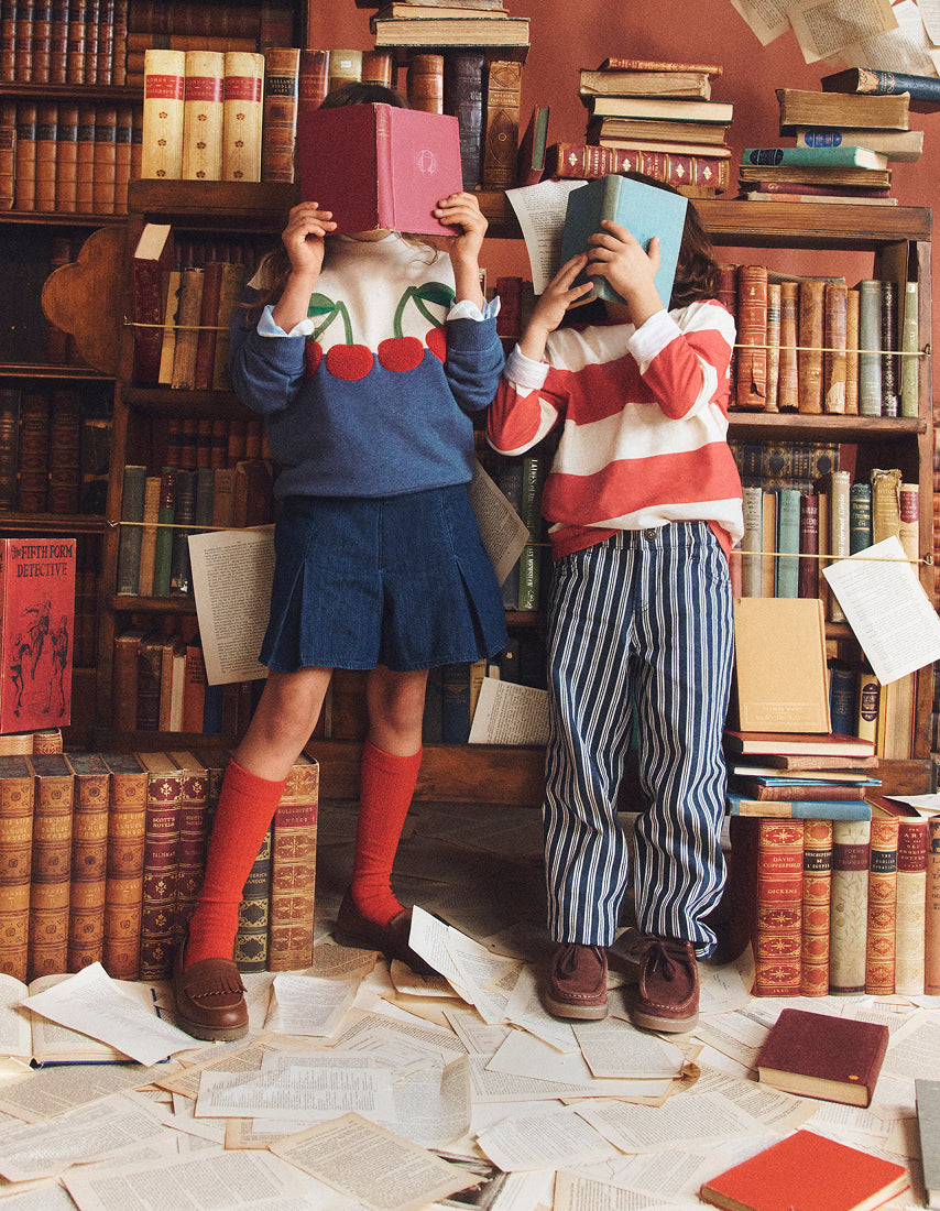 Two children standing in a book-filled room, each holding a book over their face. The child on the left wears a blue sweater with a cherry motif, a blue pleated skirt, and red knee-high socks.