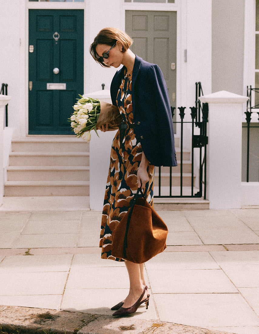 A woman stands on a sidewalk in front of a white building with dark green and gray doors. She is wearing a navy blue blazer over a brown and beige patterned midi dress, holding a bouquet of white flowers and carrying a large brown suede bag.