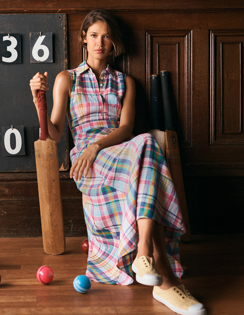 Woman in a plaid dress holding a cricket bat with sports equipment on a wooden floor.