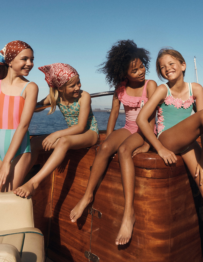 Four young girls in swimsuits sitting on a boat with a clear blue sky and ocean background.