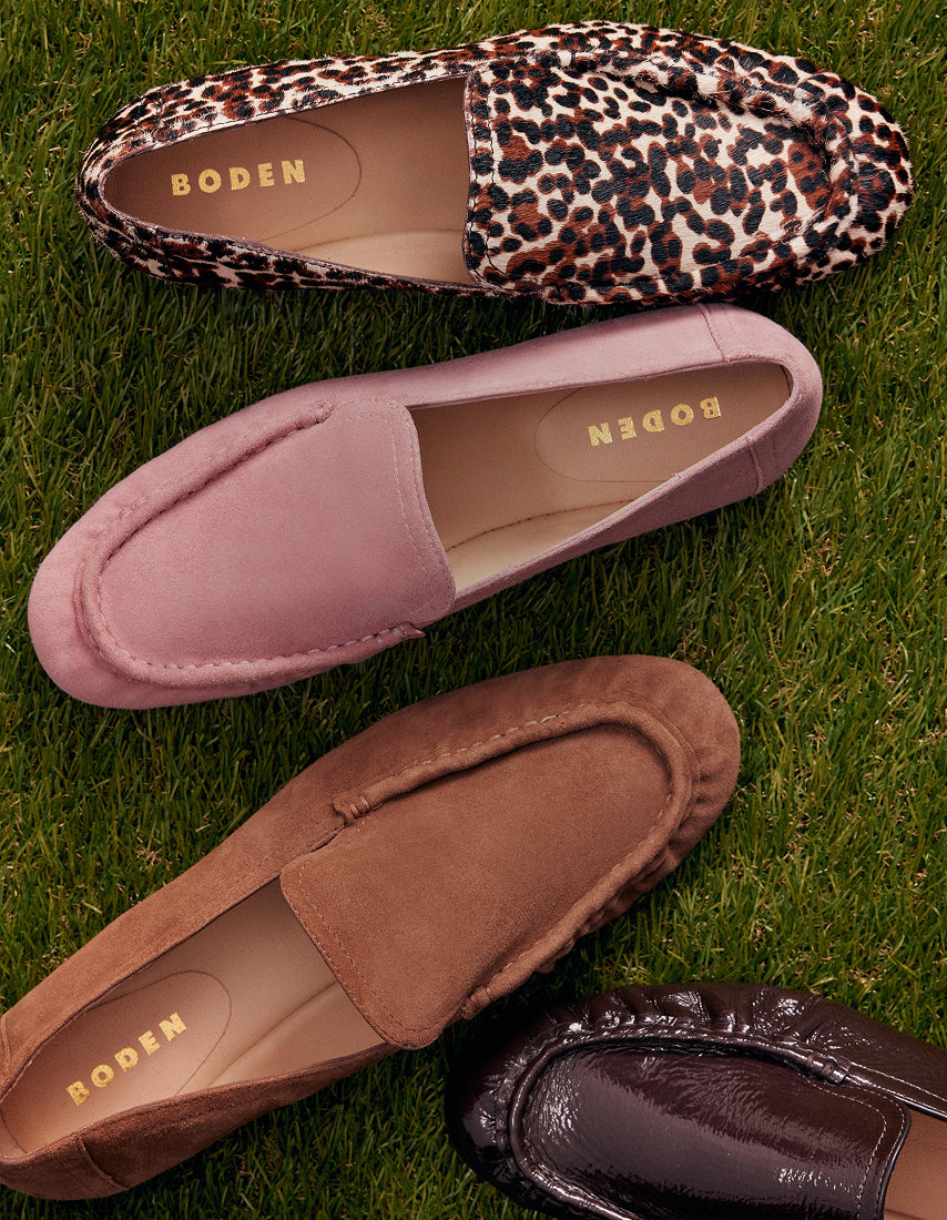 Four BODEN brand slip-on loafers arranged on green grass: a leopard print loafer, a dusty pink suede loafer, a brown suede loafer, and a dark brown leather loafer partially visible in the corner.