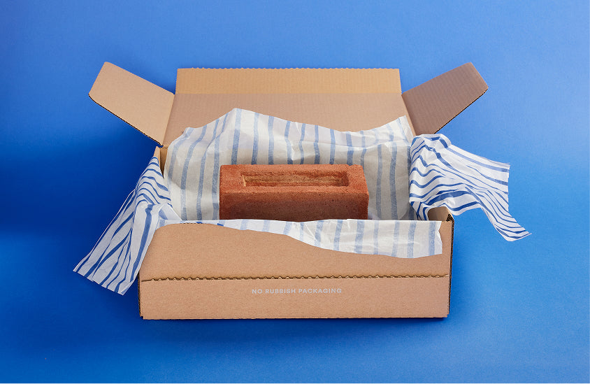 An open brown cardboard box lined with blue and white striped tissue paper, containing a rectangular terracotta-colored brick or stone block, photographed against a solid blue background.