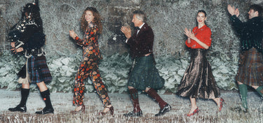 A group of five people outdoors in winter clothing, including kilts, a red top, and patterned outfits, posed against a snowy stone wall.