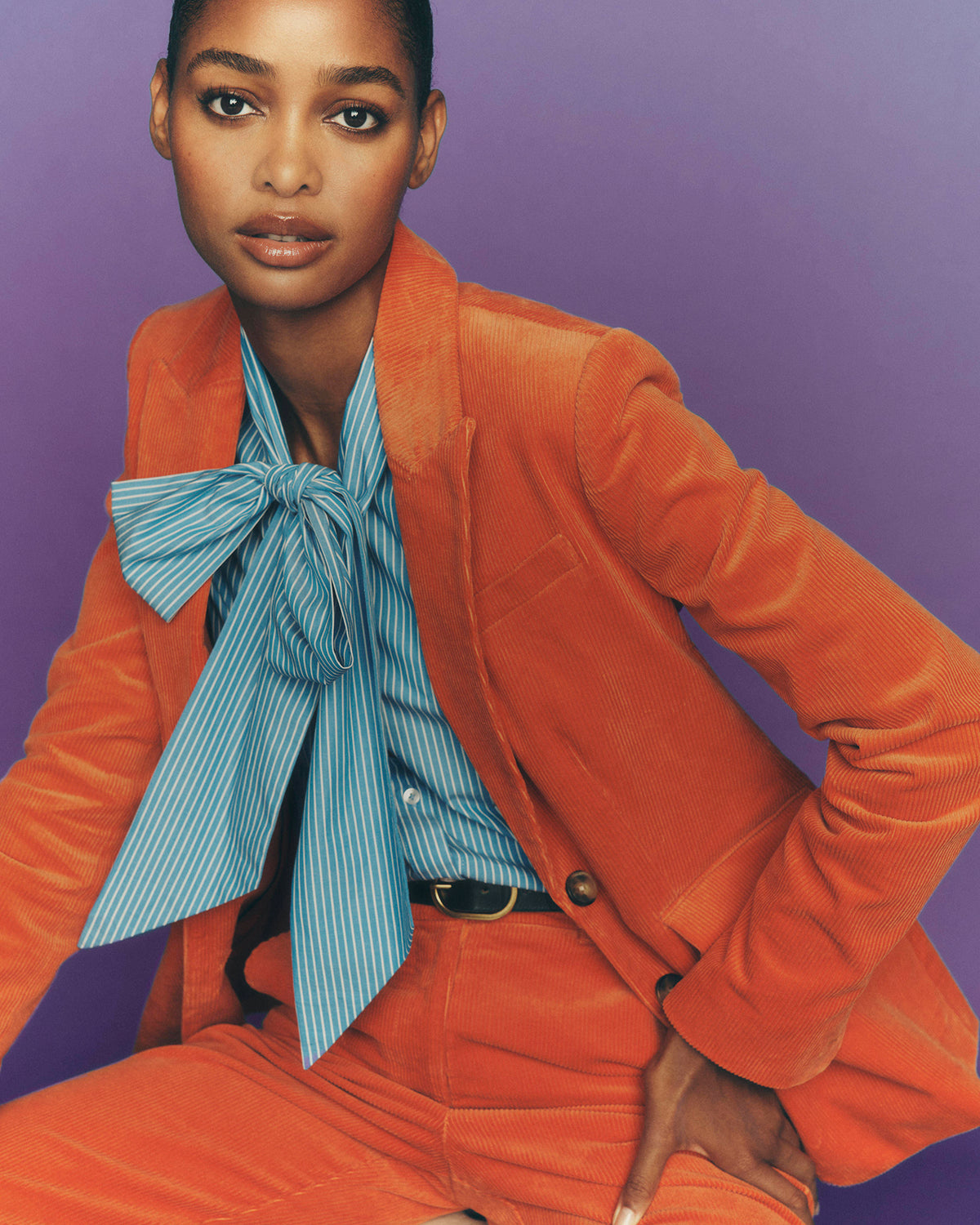 A model wearing a vibrant orange corduroy blazer and matching trousers, styled with a light blue and white striped shirt featuring a large bow tie, set against a solid purple background.
