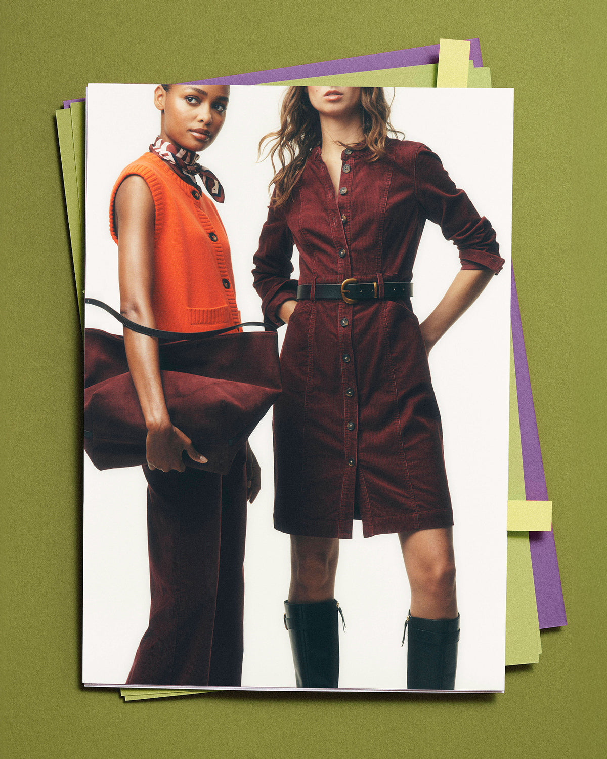 A fashion magazine or catalog page featuring two models, one in an orange vest and brown trousers holding a brown bag, and the other in a dark red corduroy shirt dress with a black belt and black boots. The page is displayed on top of several colorful paper sheets against a solid olive green background.