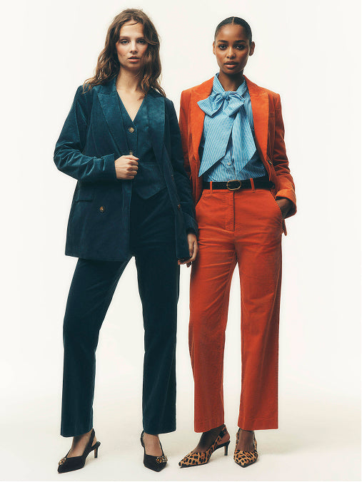 Two models standing side by side in studio fashion photography against a plain white background. The model on the left wears a dark teal velvet blazer and matching trousers with black heels.