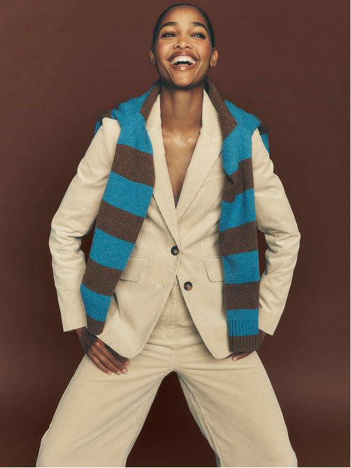 A smiling model wearing a cream-colored blazer and matching trousers, styled with a blue and brown striped knit sweater vest, posed against a solid dark brown background.