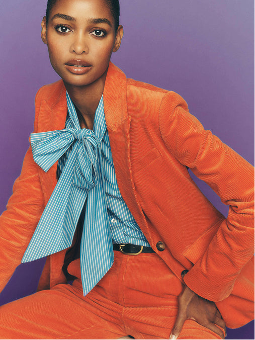 A model wearing a vibrant orange corduroy blazer and matching trousers, styled with a light blue and white striped shirt featuring a large bow tie, set against a solid purple background.