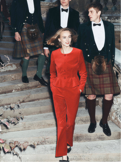 A woman in a bright red tailored suit walks down stone stairs while three men in black tuxedos and tartan kilts stand behind her.